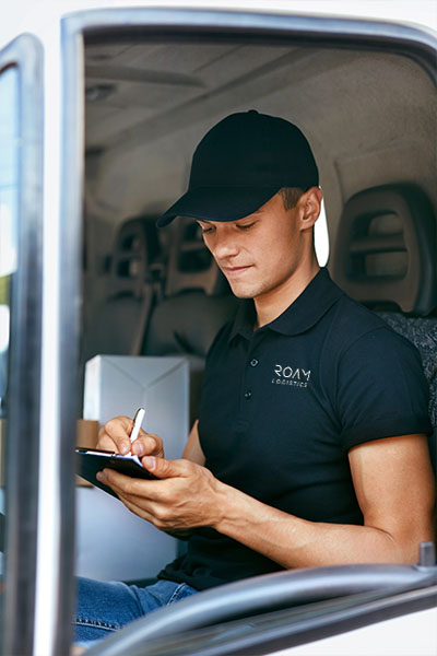 A young man is taking notes on a clipboard while sitting in his semi truck. He is wearing a blue hat and blue polo adorned with "Roam Logistics".