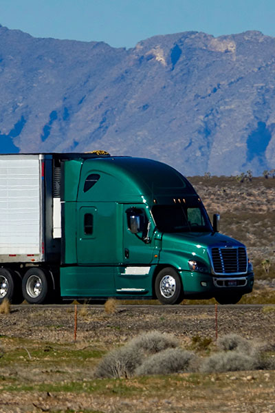 A green semi truck is driving down the highwighy with mountains in the background.