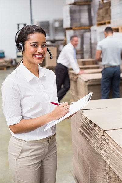Woman wearing a headset is taking notes on a clipboard while standing in a warehouse full of boxes.